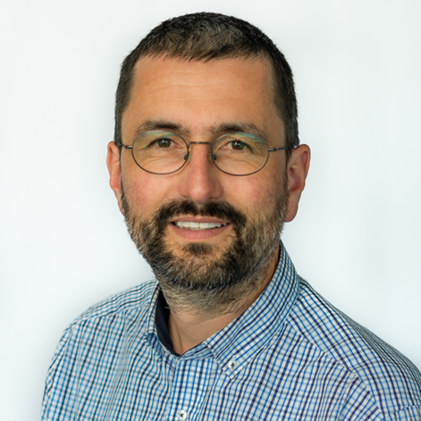 Ein Mann mit kurzem dunklem Haar, Brille und Vollbart trägt ein kariertes Hemd. Er lächelt leicht und steht vor einem hellen, neutralen Hintergrund.