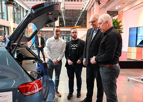 Vier Männer stehen an einem geöffneten Kofferraum eines Autos in einem modernen, hellen Gebäude. Sie schauen gemeinsam in den Kofferraum, in dem technische Geräte eingebaut sind. Im Hintergrund sind weitere Personen und eine bunte Wand zu sehen.