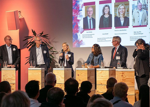 Sechs Personen, drei Männer und drei Frauen, stehen und sitzen hinter erhöhten Rednerpulten auf einer Bühne bei einer Podiumsdiskussion. Über ihnen ist eine große Leinwand mit Porträtfotos und Namen der Teilnehmenden. Das Publikum sitzt im Vordergrund und hört zu.