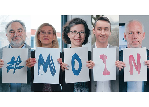 Fünf Menschen stehen nebeneinander und halten jeweils ein Schild mit einem Buchstaben oder Symbol, sodass zusammen „#MOIN“ zu lesen ist. Alle blicken freundlich in die Kamera.