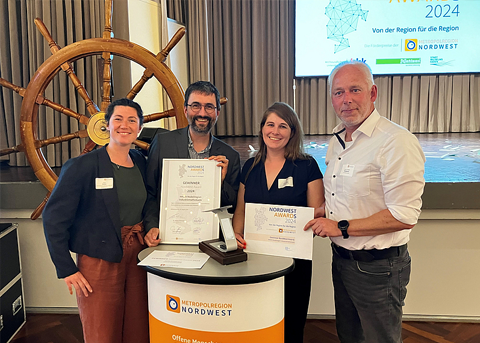 Zwei Frauen und zwei Männer stehen lächelnd an einem Stehtisch in einem Veranstaltungssaal und halten eine Urkunde und einen Award in der Hand. Hinter ihnen ist ein großes Steuerrad und eine Leinwand mit dem Schriftzug „Nordwest Award 2024“.
