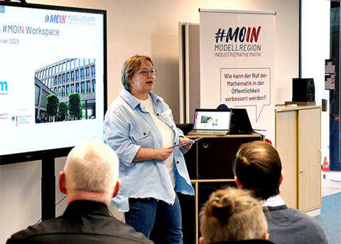 Eine Frau hält einen Vortrag vor sitzendem Publikum. Sie steht neben einem großen Bildschirm mit der Aufschrift „#MOIN Workspace“ und einem Foto eines Gebäudes. Rechts daneben steht ein Banner mit dem Slogan „#MOIN Modellregion Industriemathematik“.