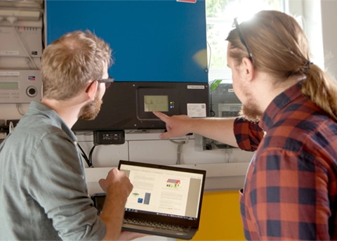 Zwei Männer analysieren Energiedaten an einem Gerät und Laptop in einem technischen Umfeld.